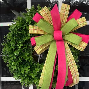 May include: A circular green wreath adorned with a colourful bow. The bow incorporates bright pink, green, and yellow plaid ribbons. The wreath is composed of small, closely packed green leaves, giving it a full and textured look. The bow is the central feature.