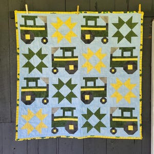 Op de afbeelding: Een handgemaakte quilt met een lichtblauwe achtergrond heeft herhaalde patronen van groene tractoren, gele sterren en donkergroene sterren. De quilt is afgezet met een geel en groen patroon en wordt opgehangen met houten wasknijpers.
