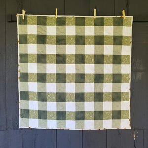 May include: A square quilt with a green and white checkered pattern, hanging from wooden clothespins. The quilt is made of fabric with a repeating pattern of squares. The background is a dark gray wooden wall.