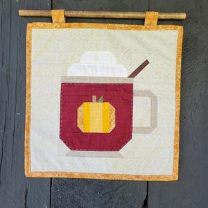 May include: A quilted wall hanging featuring a mug of pumpkin spice latte. The mug is burgundy with a yellow pumpkin applique. The drink has white whipped cream and a brown stir stick. The wall hanging is square with an orange border and a wooden dowel.