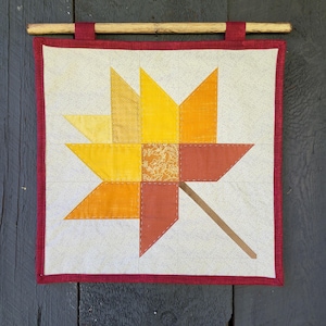 May include: A handmade wall hanging featuring a stylized leaf design. The leaf is composed of various shades of yellow, orange, and brown fabric, with a brown stem. The quilt is framed with a red border and hung from a wooden dowel.