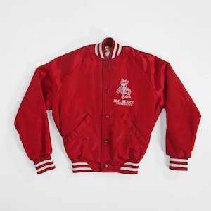 Op de afbeelding: Een rode satijnen bomberjack met wit en rood gestreepte manchetten en kraag. De jas heeft een wit geborduurd logo van een wolfachtig mascotte en de tekst "N.C. State University". De jas heeft een knoopsluiting en twee voorzakken.