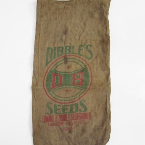 Puede incluir: Un saco de semillas de yute vintage con el texto "DIBBLE'S SEEDS" en verde y rojo. El saco presenta un logotipo circular con "DB" en su interior. El texto "EDWARD E. DIBBLE SEEDGROWER HONEOYE FALLS N.Y." está impreso debajo.