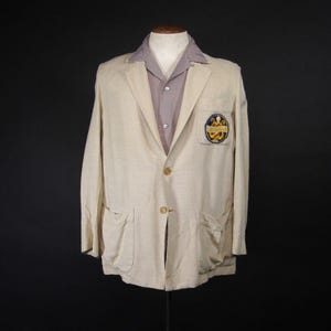May include: A beige blazer with a patch on the left pocket. The patch is a circle with a gold design and the text "The Golden Gate" in blue. The blazer has two pockets on the front and a button closure.