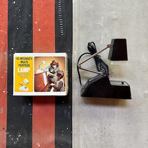 May include: A black and white high-intensity multi-purpose lamp with an adjustable metal arm and a rectangular base. Next to it is a cardboard box featuring the text HI-INTENSITY MULTI PURPOSE LAMP and an illustration of a person playing a piano.