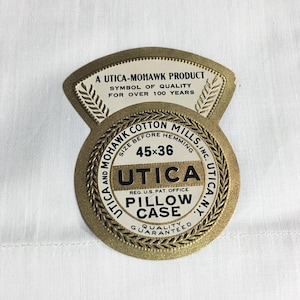 May include: A gold label with the text "A Utica-Mohawk Product, Symbol of Quality for Over 100 Years" and "Utica and Mohawk Cotton Mills, Inc. Utica, N.Y."  The label also includes the text "Size Before Hemming 45x36", "Utica", "Reg. U.S. Pat. Office", "Pillow Case", and "Quality Guaranteed".