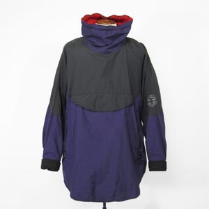 May include: A dark blue and black anorak jacket with a red lining around the collar. The jacket has a zipper closure and a large pocket on the front.