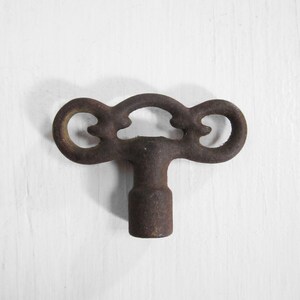 May include: A rusty metal key with a decorative, ornate head. The key has a T-shaped handle and two loops on either side of the head.