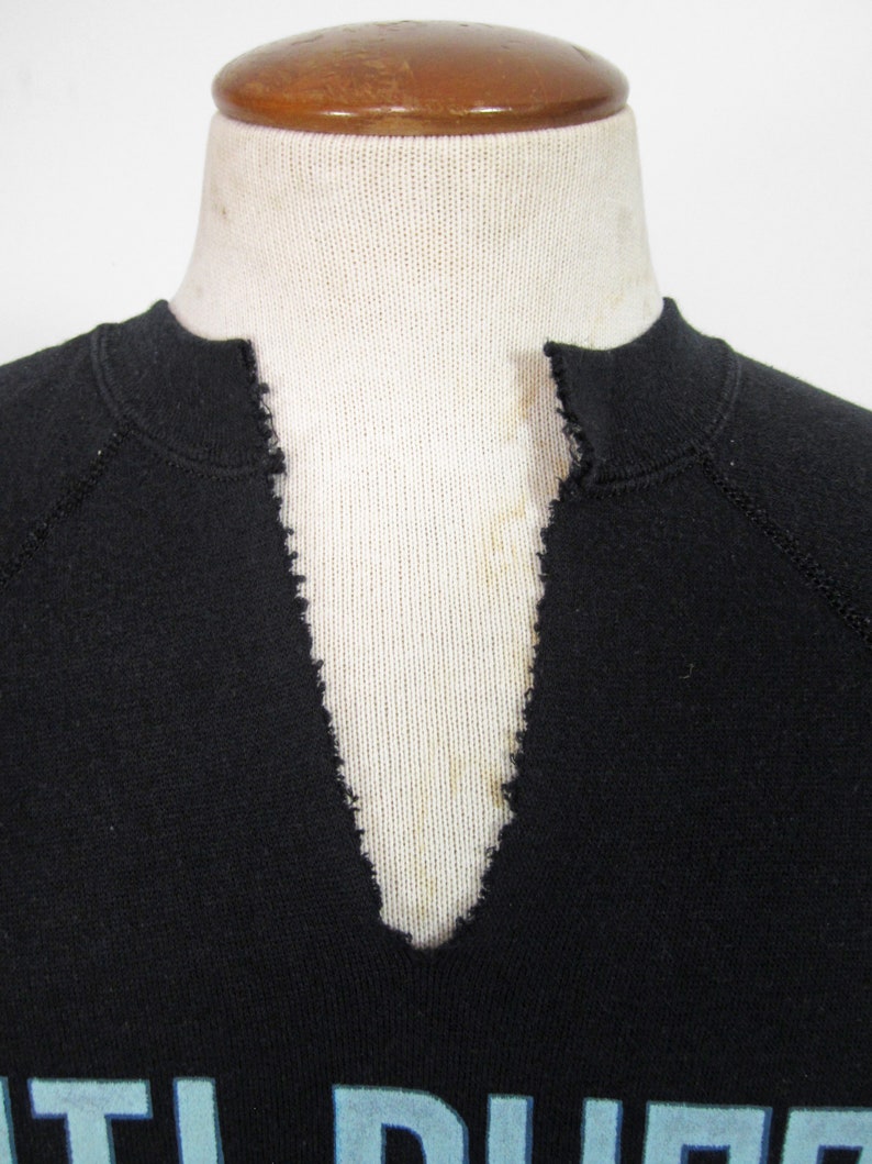 May include: Close-up of the neckline of a black sweatshirt with a V-neck. The neckline is frayed and has a light blue stain.