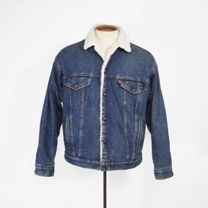 May include: A blue denim jacket with a white, fluffy shearling lining and collar. The jacket has two chest pockets, two side pockets, and a button-up front. A small red tag is visible on the chest pocket.