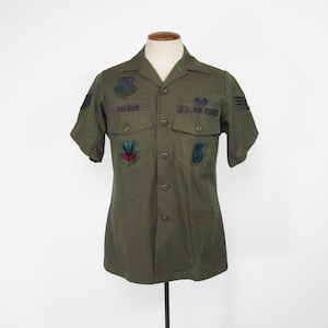 May include: Olive green U.S. Air Force short-sleeved shirt with various patches and the name "SALMAN" above the left pocket. The shirt features two chest pockets and is buttoned. It is displayed on a mannequin.