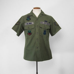 Peut inclure: Chemise vert olive à manches courtes avec plusieurs écussons et les inscriptions "SALMAN" et "U.S. AIR FORCE". La chemise boutonnée est présentée sur un mannequin. Les écussons comprennent des emblèmes et des insignes.