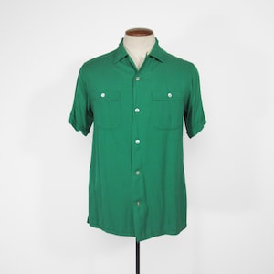 May include: A short-sleeved, emerald green shirt with two chest pockets and a button-down front. The shirt is displayed on a mannequin, showcasing its classic design and tailored fit. The shirt's color is vibrant and eye-catching.