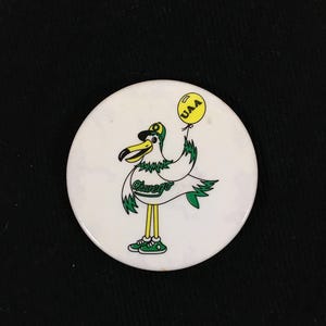 May include: A white circular pin featuring a cartoon seagull wearing a baseball cap and holding a yellow balloon with "UAA" on it. The seagull has green and white feathers and the word "Oswego" is written on the pin.