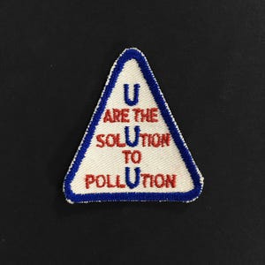Könnte beinhalten: Ein dreieckiger Aufnäher mit weißem Hintergrund und blauem Rand. Der Aufnäher zeigt den Text "U ARE THE SOLUTION TO POLLUTION" in roter und blauer Schrift.