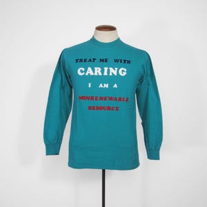 Może przedstawiać: Turkusowa koszulka z długim rękawem z napisem "TREAT ME WITH CARING I AM A NONRENEWABLE RESOURCE" w białych i czerwonych literach. Koszulka jest prezentowana na manekinie na białym tle.