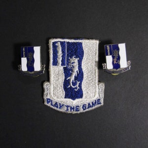 Pode incluir: Um conjunto de três patches militares com um brasão azul e branco com um design de dragão. O patch maior tem o texto "Play the Game" abaixo do brasão. Os patches menores têm o texto "Play the Game" acima do brasão.