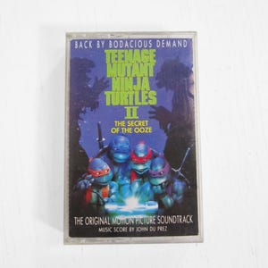 Könnte beinhalten: Eine Kassette von Teenage Mutant Ninja Turtles II: Das Geheimnis des Ooze. Das Cover zeigt die vier Schildkröten mit ihren farbigen Masken und dem Text "BACK BY BODACIOUS DEMAND". Der Original-Soundtrack des Films ist ebenfalls aufgeführt.
