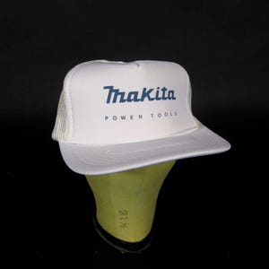 Op de afbeelding: Een witte truckerpet met een schuimrubberen voorpaneel voorzien van het blauwe Makita-logo en de tekst POWER TOOLS eronder gedrukt. De pet heeft een wit gaas aan de achterkant en een platte klep, met een strak ontwerp en een gestructureerde kroon.