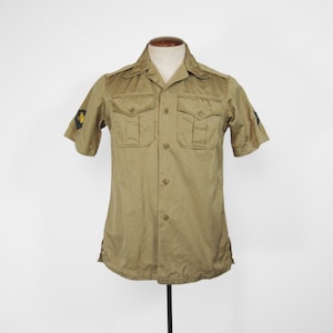 May include: A khaki-coloured short-sleeved shirt with two chest pockets and button fastenings. The shirt has epaulettes on the shoulders and patches on the sleeves. The shirt is displayed on a mannequin against a white background.