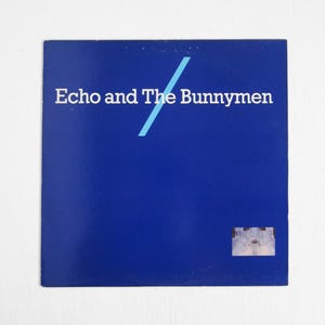 May include: A square, blue album cover with the text "Echo and The Bunnymen" in white. A light blue diagonal line bisects the text. A small, rectangular image of a building is in the lower right corner.