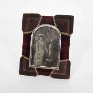 May include: An antique photo frame with a silver-colored metal frame holding a black and white photograph of a couple. The frame is set against a burgundy velvet background with brown velvet accents and gold stitching.