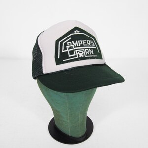 May include: A trucker hat with a white front panel and a black mesh back. The hat features a dark green brim and a graphic on the front that reads "Pampers Barn" in white and dark green.
