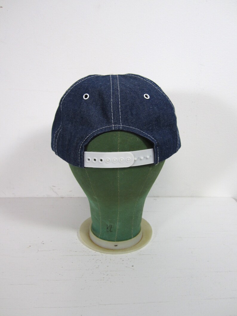 Puede incluir: Una gorra de b&eacute;isbol de mezclilla azul con una correa ajustable de pl&aacute;stico blanco. La gorra est&aacute; en una cabeza de maniqu&iacute; verde.