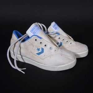 Puede incluir: Un par de zapatillas de lona blancas vintage con detalles y cordones azules. Las zapatillas presentan el logotipo de la estrella Converse y el texto "CHRIS EVERT CONVERSE" en la lengüeta. Las zapatillas están sobre un fondo negro.