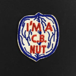 Puede incluir: Un parche blanco bordado con costuras azules y rojas. El parche tiene forma de nuez y dice "I'M A C.B. NUT".
