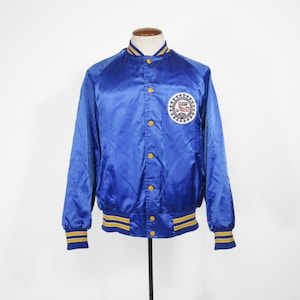 May include: A blue satin bomber jacket with yellow and blue striped cuffs and waistband. The jacket has a large circular patch on the left chest with the text "E.A.S.W. 1978" inside.