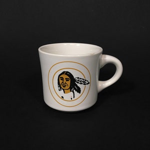 Könnte beinhalten: Weiße Keramiktasse mit einer schwarzen und goldenen Illustration eines Indianers mit Federkopfschmuck. Die Illustration ist von einem goldenen Kreis umgeben.