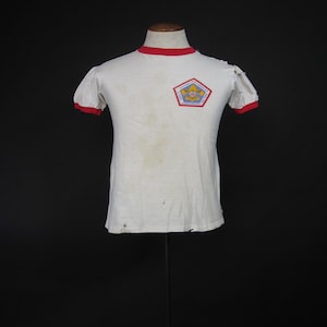 Könnte beinhalten: Weißes T-Shirt mit rotem Besatz am Hals und an den Ärmeln. Das Shirt hat einen Boy Scouts of America-Aufnäher auf der Vorderseite mit dem Text "75th Anniversary 1910-1985".