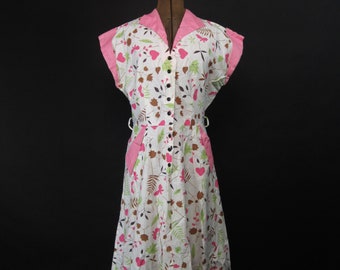 Puede incluir: Un vestido blanco con cuello rosa y estampado floral de hojas y corazones. El vestido tiene un cierre de botones en la parte delantera y bolsillos en los lados.