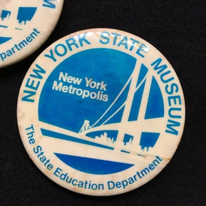 May include: Two vintage round buttons with a blue and white design. The buttons feature the text "NEW YORK STATE MUSEUM" and "New York Metropolis". The bottom of the button reads "The State Education Department".