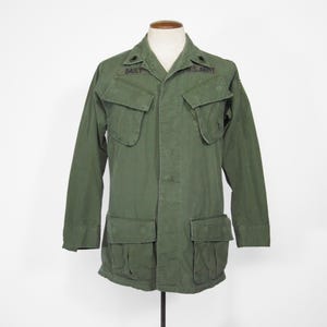 Puede incluir: Chaqueta militar verde oliva con cuatro bolsillos delanteros y cuello. La chaqueta tiene el nombre "BAILY" encima del bolsillo izquierdo y "U.S. ARMY" encima del bolsillo derecho. La chaqueta se exhibe en un maniquí.