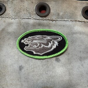 Op de afbeelding: Een lichtgrijze canvasstof met drie donkerbruine metalen ringen langs de bovenrand. In het midden van de stof bevindt zich een ovaal geborduurde patch met een fel limoengroene rand, een zwarte achtergrond en een grijze grommende panterkop.