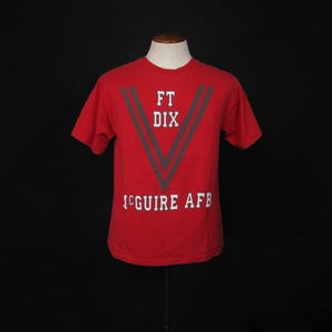 May include: Red t-shirt with a grey and white graphic design. The graphic features a large V shape with the text "FT DIX MCGUIRE AFB" inside.