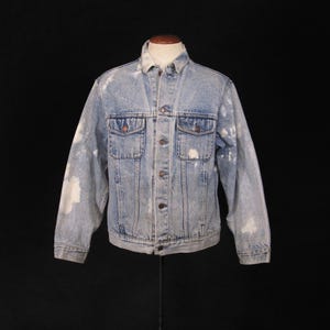 May include: A light blue denim jacket with white bleach splatters. The jacket has a button-up front, two chest pockets, and long sleeves.