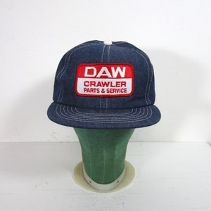 Puede incluir: Un gorro de camionero de mezclilla azul con una malla blanca en la parte posterior. El gorro tiene un parche rojo y blanco que dice "DAW CRAWLER PARTS & SERVICE".