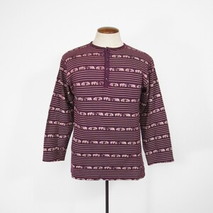 May include: A long-sleeved shirt with a purple background and a pattern of white elephants. The shirt has a button-up collar and a single button on the front.