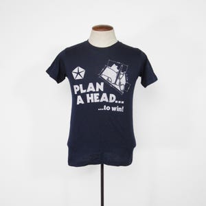 Pode incluir: Uma camiseta azul marinho com um gráfico branco de um carro e o texto "PLAN A HEAD... ...to win!"