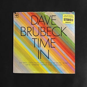 May include: A vintage vinyl record album cover for "Time In" by Dave Brubeck. The cover features a colorful diagonal stripe design in shades of orange, yellow, blue, and green. The album title and artist name are in bold black text.