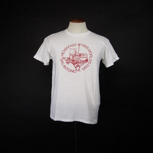 May include: White t-shirt with a red print of the 1968 World's Fair HemisFair in San Antonio, Texas. The print features a stylised outline of the state of Texas with the fair's logo and buildings.