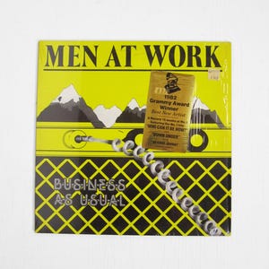 May include: A vinyl record album cover for the band Men at Work, titled "Business as Usual." The cover is yellow with black and white graphics, including mountains and a telephone cord. It also features a Grammy Award sticker.