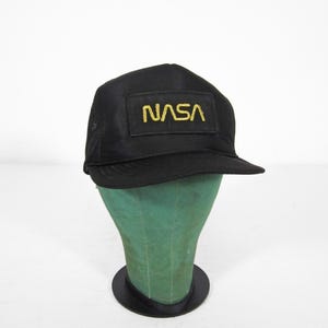 Könnte beinhalten: Schwarze Baseballkappe mit dem NASA-Logo in goldfarbenen Buchstaben. Die Kappe hat einen gebogenen Schirm und wird auf einem grünen Schaufensterkopf präsentiert. Die Kappe besteht aus einem dunklen Material, möglicherweise Nylon.