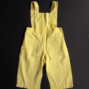Könnte beinhalten: Hellgelbe Kinder-Latzhose mit verstellbaren Trägern und Knopfverschlüssen. Das Kleidungsstück hat ein klassisches Latzdesign mit kurzen Beinen und zwei kleinen Taschen. Der Stoff scheint eine leichte Baumwoll- oder Leinenmischung zu sein.