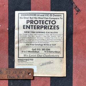 May include: A vintage Protecto Enterprizes New 1984 Spring Catalog for Commodore 64 and VIC-20 owners. The paper catalog features black text on a light background, including contact information and a wooden ruler showing a 9 cm segment.