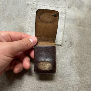 May include: A small, rectangular, dark brown leather case with a hinged flap. The exterior features a textured, weathered finish and a centered oval cutout on the lower section, revealing a light-colored, fuzzy interior lining material.