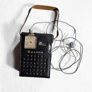 May include: Vintage Bulova watch with a black leather case and a brown strap. The watch has a rectangular face with a light-colored dial and gold-tone hands. The case has a series of small holes and the word "Bulova" is printed on the case.
